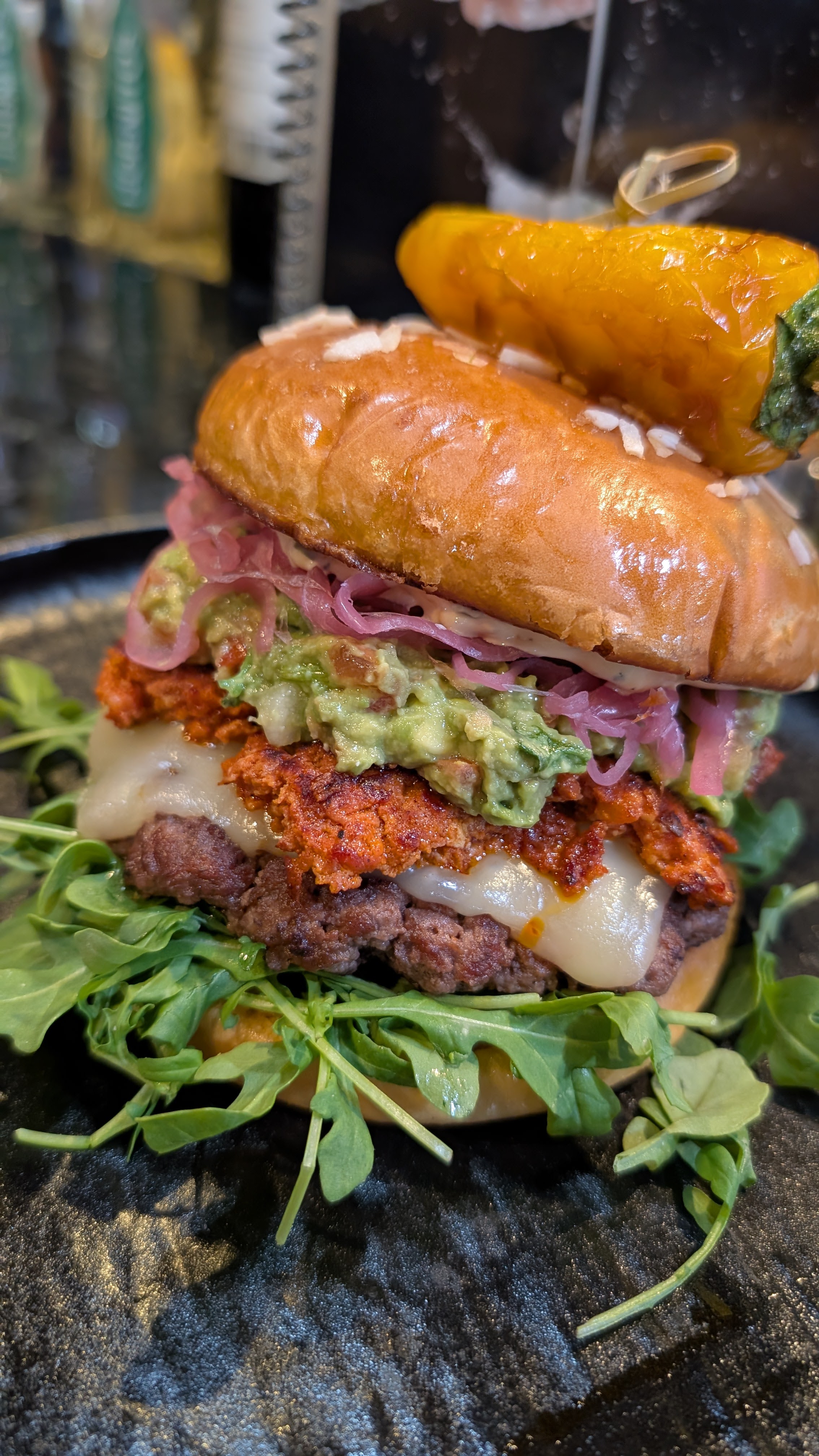 A stacked craft burger with guacamole, pickled onions, arugula, and a grilled pepper.